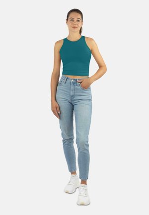 Junge Frau steht mit einer Hand in der Tasche, trägt ein aquafarbenes ärmelloses Crop-Top, hellblaue High-Waist-Jeans und weiße Sneaker vor einem weißen Hintergrund.