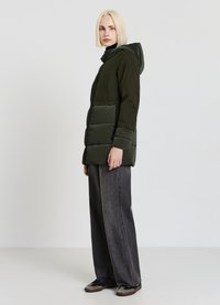 Giacca imbottita verde scuro con cappuccio, con una texture trapuntata nella metà inferiore, abbinata a jeans grigi a gamba larga e scarpe nere slip-on.