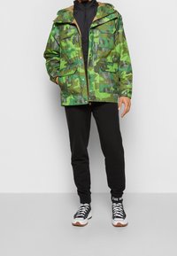 Green camouflage jacket with a hood, front pockets, and a shiny finish, paired with black sweatpants and black high-top sneakers.