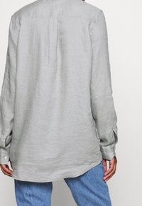 Camisa de lino gris claro con mangas largas, dobladillo redondeado y un pliegue en la parte trasera, combinada con jeans de denim. Tejido texturizado y transpirable.