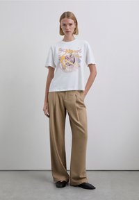 White graphic t-shirt with colorful print featuring a bird and text. Paired with loose beige trousers and black slip-on shoes. Neutral background.