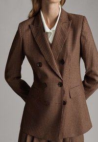 Brązowa, kratkowana marynarka dwurzędowa z wyraźną strukturą, klapami w szpic i dużymi czarnymi guzikami. Teksturowany materiał z kieszeniami.