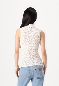 Čipkast bezrukavni top u off-white boji, s cvjetnim uzorcima i visokim ovratnikom. Prozirni dizajn se savršeno uklapa s ležernim trapericama.