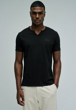 Joven hombre con una camiseta negra ajustada de manga corta y pantalones beige claros, de pie contra un fondo gris liso con las manos relajadas.