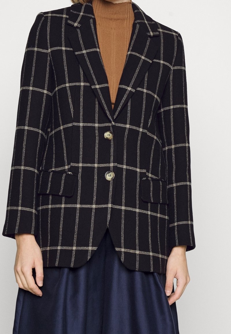 Femme portant un blazer noir à carreaux blancs, un col roulé beige et une jupe bleu marine foncé sur un fond uni.