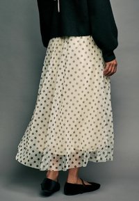 Pull noir, jupe crème avec un superposé en tulle à pois, design fluide, longueur mollet, et chaussures plates noires. Texture lisse et motif ludique.