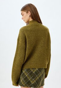 Oliwkowy sweter dzianinowy z wysokim kołnierzem, luźny krój i teksturowany materiał, zestawiony z plisowaną spódnicą w zielono-żółte wzory.