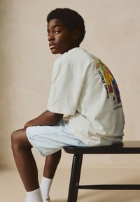 White short-sleeve t-shirt with vibrant graphic print on the back, paired with light blue denim shorts. Sitting on a black chair.