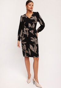 Czarna sukienka z długimi rękawami i dekoltem w serek, zdobiona białym wzorem liści. Podkreślona talia za pomocą paska i gładka tekstura.