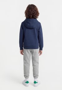 Barn med krøllete hår står med ryggen til, iført en marineblå hettegenser, lysegrå joggebukser, hvite sokker og hvite sneakers med grønne detaljer.