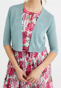 Světlounce modrý krátký kardigan s krátkými rukávy, vyrobený z lesklého materiálu, nošený přes šaty s květinovým potiskem v růžových a červených tónech.