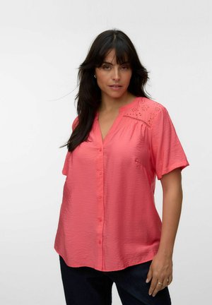 Frau mit dunklen Haaren, die eine korallfarbene, kurzärmelige Bluse mit Knöpfen und Spitzendetails an den Schultern trägt, steht vor einem weißen Hintergrund.