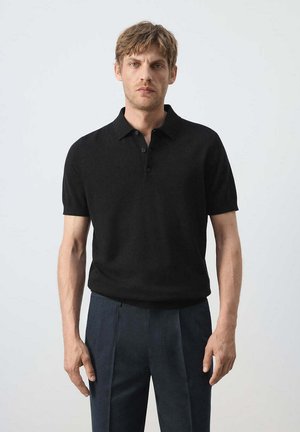 Hombre con cabello corto castaño claro, que lleva una camisa tipo polo negra de manga corta y pantalones plisados azul oscuro, de pie contra un fondo liso.