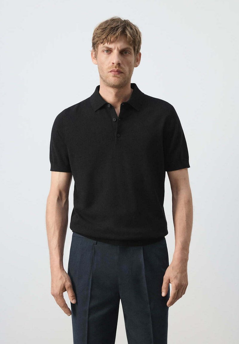 Homme aux cheveux courts châtain clair, portant un polo noir à manches courtes et un pantalon plissé bleu foncé, debout devant un fond uni.