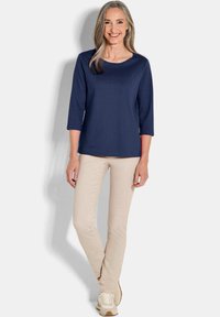 Marineblaues Oberteil mit U-Ausschnitt und Dreiviertelärmeln; kombiniert mit hellbeigen, schmal geschnittenen Hosen und weißen Sneakers. Glatte Stofftextur.