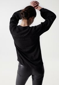 Maglione nero a maniche lunghe con tessuto strutturato e dettaglio arricciato sul retro, abbinato a pantaloni neri aderenti con tasche decorative.