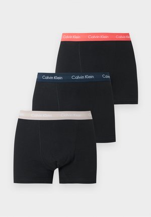 Szczupły ciemnoskóry mężczyzna stoi boso, ubrany w czarne bokserki Calvin Klein na jednolitym jasnym tle.