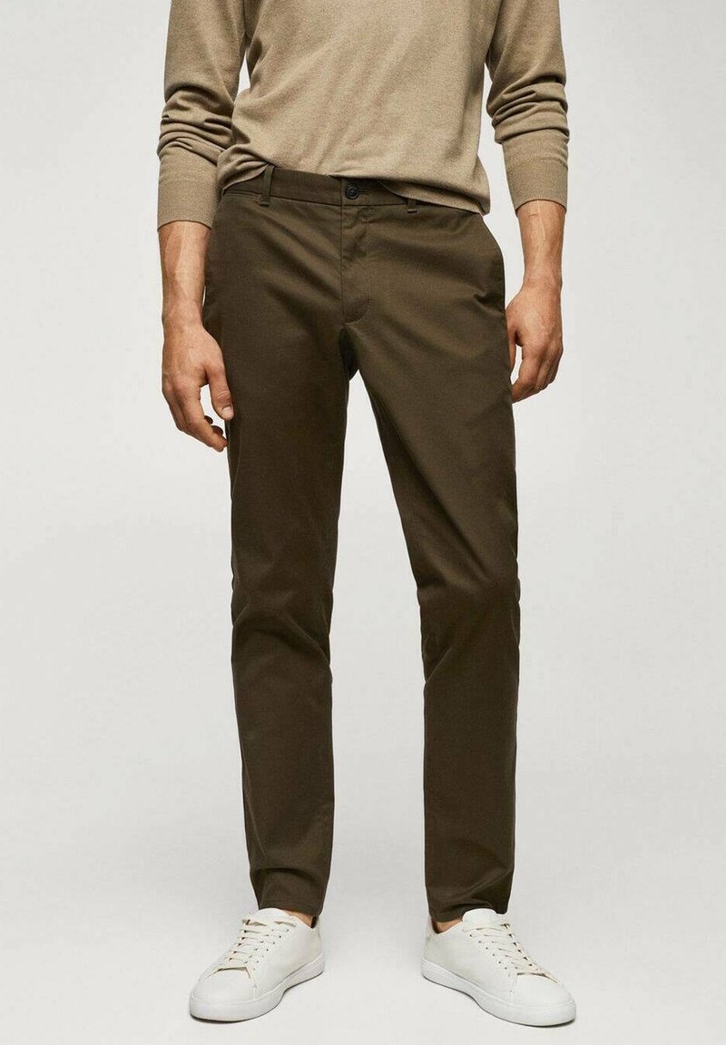 Homme portant un pantalon slim marron foncé, une chemise beige à manches longues et des baskets blanches, debout les mains détendues le long du corps.