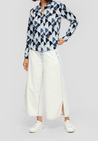 Button-down-Hemd mit geometrischem Muster in Blautönen, kombiniert mit weit geschnittenen weißen Denim-Hosen mit seitlichen Schlitzen. Weiße Sneakers.