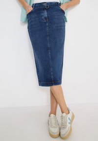 Personne portant une jupe en jean bleu foncé longueur genou avec ceinture et des baskets blanches à accents bleus, debout les jambes croisées.