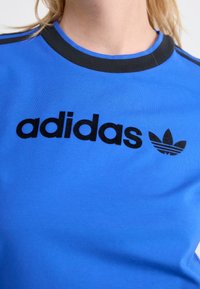 Blå bomull t-skjorte med svart "adidas" logo og emblem, rund svart krage og kontrasterende svarte striper på skuldrene.