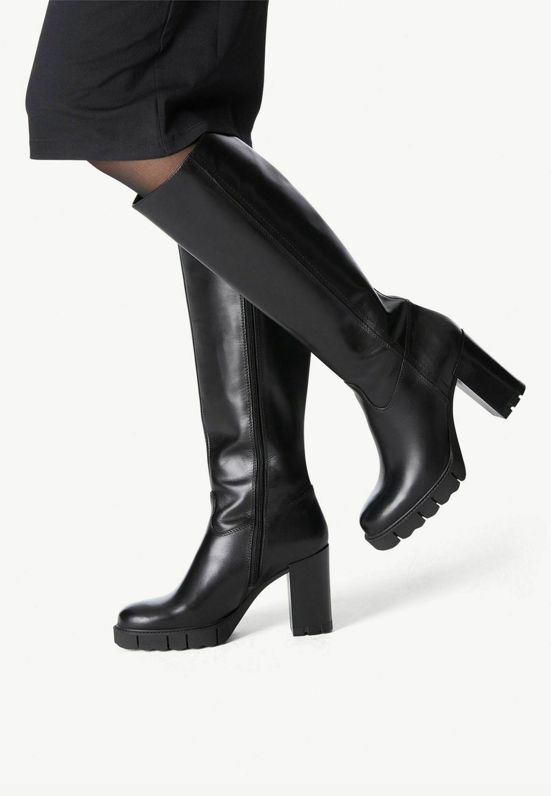 Botas de cuero negro hasta la rodilla, fabricadas en piel suave. Presentan un tacón grueso y una suela texturizada y estriada. Detalle de cremallera en el lateral.