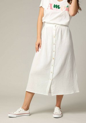 Jupe midi blanche en tissu texturé avec une fermeture à boutons sur le devant. Associée à un t-shirt graphique blanc et des baskets blanches, montrant un style décontracté.