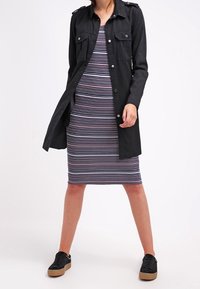 Veste chemise longue noire avec poches portée sur une robe ajustée rayée bleu marine, blanche et rouge. Baskets noires avec une semelle texturée.