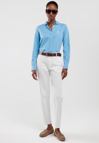 Lichtblauw gestreept overhemd met lange mouwen, witte broek, bruine riem en tan loafers. Zwarte zonnebril maakt de outfit af.