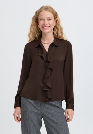 Femme souriante aux cheveux blonds bouclés portant un chemisier marron foncé à volants et un pantalon gris, debout devant un fond clair uni.