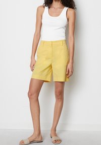 Vrouw in een witte top, gele kniebandschoenen met zakken en beige slipper sandalen, staand tegen een witte muur.