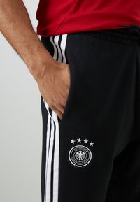 Calças de moletom pretas com riscas brancas nas laterais, apresentando um logótipo circular com cinco estrelas e o texto "Deutscher Fußball-Bund."