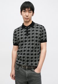 Čierne polokošeľa s geometrickým bielym vzorom, krátkymi rukávmi a čiernym golierom. Nosené s tmavosivými džínsy a hnedým opaskom.