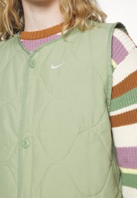 Person som bär en vadderad ljusgrön Nike-väst över en flerfärgad randig stickad tröja med knappar på västens framsida.