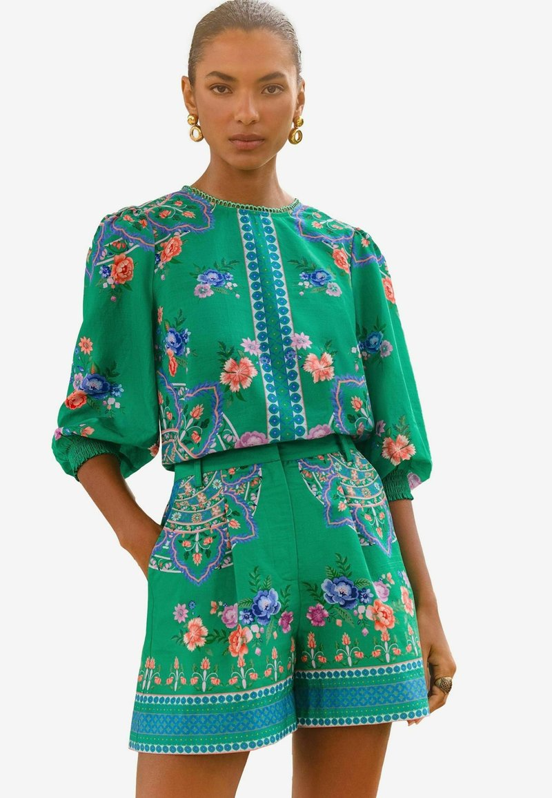 Zaļa ziedu divdaļīga tērpa, kas izceļas ar sarežģītu izšuvumu, puffed piedurknēm un dekoratīviem rakstiem ar ziliem un rozā akcentiem.