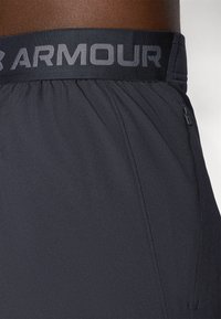Schwarze Sportshorts verfügen über einen elastischen Bund mit der Aufschrift "ARMOUR" in grauen Buchstaben, strukturiertem Stoff und einer seitlichen Reißverschlusstasche.