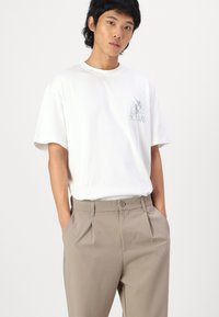 White cotton t-shirt with short sleeves and a blue graphic print, paired with light beige pleated trousers, featuring side pockets.