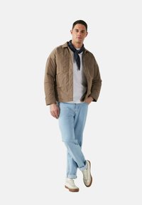 Giacca trapuntata marrone sopra una felpa grigia, jeans blu chiaro e sneakers bianche con dettagli marroni. Una sciarpa blu navy aggiunge contrasto.