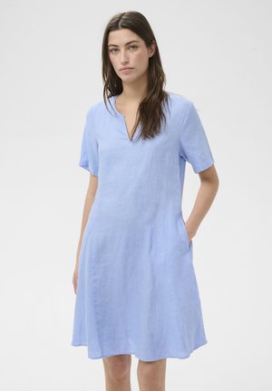 Femme aux longs cheveux bruns portant une robe bleu clair à manches courtes avec des poches, debout devant un fond blanc uni.