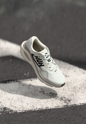 White running shoe with "RUN" text mid-air above black and white asphalt surface with dust particles around.