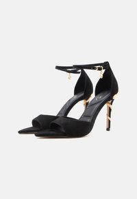 Zwarte suède sandalen met hoge hak, open teen, enkelbandje en gouden accent nabij de hiel. Voorzien van een slanke, puntige neus en een dunne stilettohak.