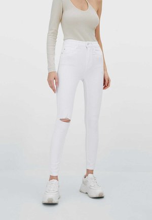 Vrouw draagt witte gescheurde skinny jeans, witte chunky sneakers en een beige asymmetrische top met lange mouwen tegen een effen achtergrond.
