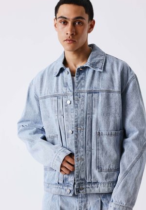 Jeune homme en veste en denim bleu clair avec poches et broderies, posant devant un fond blanc uni.