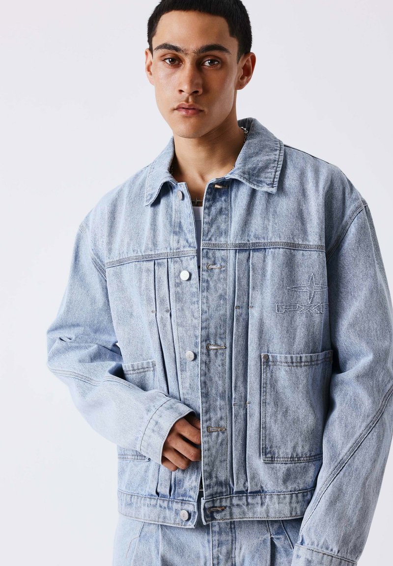 Jeune homme en veste en denim bleu clair avec poches et broderies, posant devant un fond blanc uni.