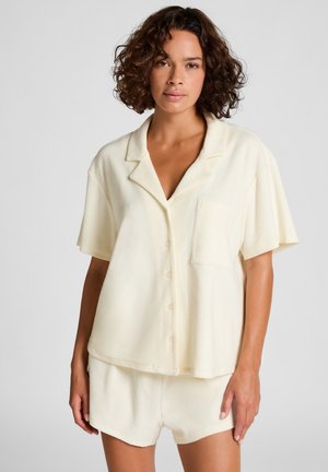 Woman with curly hair wearing a cream short-sleeve button-up shirt and matching shorts against a plain background.