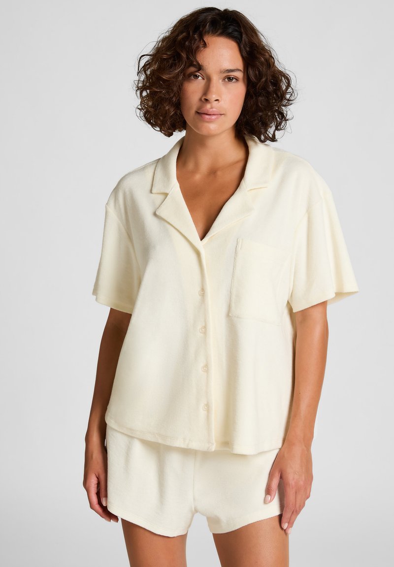 Woman with curly hair wearing a cream short-sleeve button-up shirt and matching shorts against a plain background.