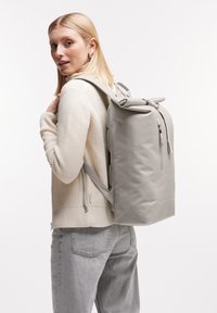 Sac à dos gris à rabat, fabriqué dans un matériau lisse, avec des bretelles réglables, une poche zippée et un design minimaliste sans logos visibles.