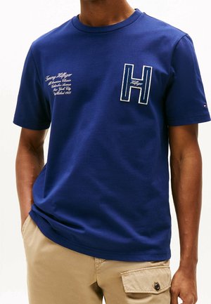 Homme portant un t-shirt bleu à manches courtes avec un texte blanc brodé et un grand "H" sur la poitrine, associé à un pantalon cargo beige.