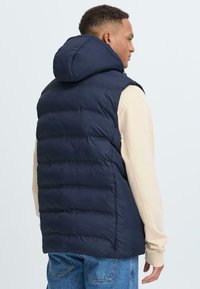 Marineblauer Steppweste mit Kapuze, ausgestattet mit gesteppten Abschnitten und einer glatten Textur, getragen über einem hellen, langärmligen Oberteil und Jeans.
