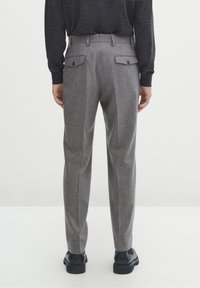 Pantalon gris sur mesure avec une texture subtile, deux poches arrière avec fermetures à boutons, et une coupe droite, assorti à des chaussures noires.